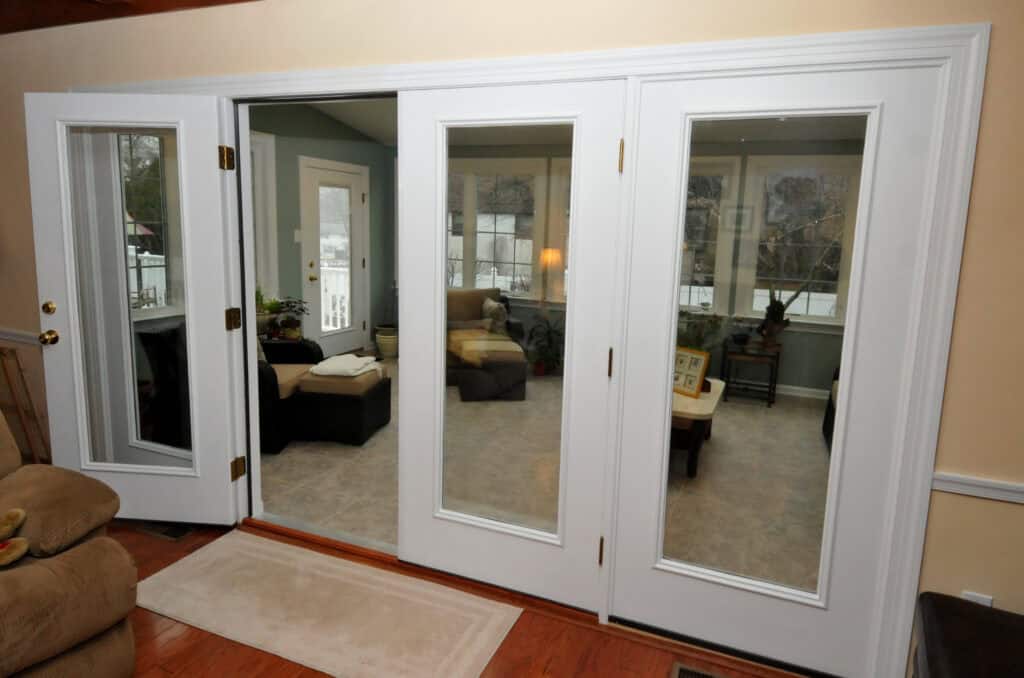 Three white-framed glass patio doors open into a sunroom, revealing comfortable seating, a small table, plants, and large windows displaying a snowy scene outside.