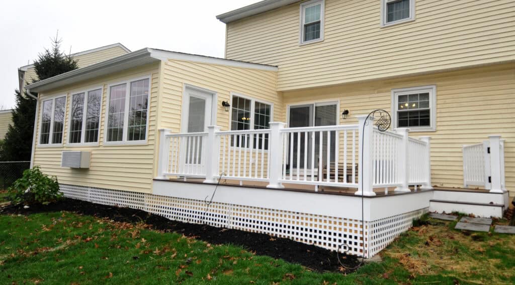 A yellow house with white trim features a sunroom with many windows and an attached white-railed deck overlooking a green yard with patches of fallen leaves.