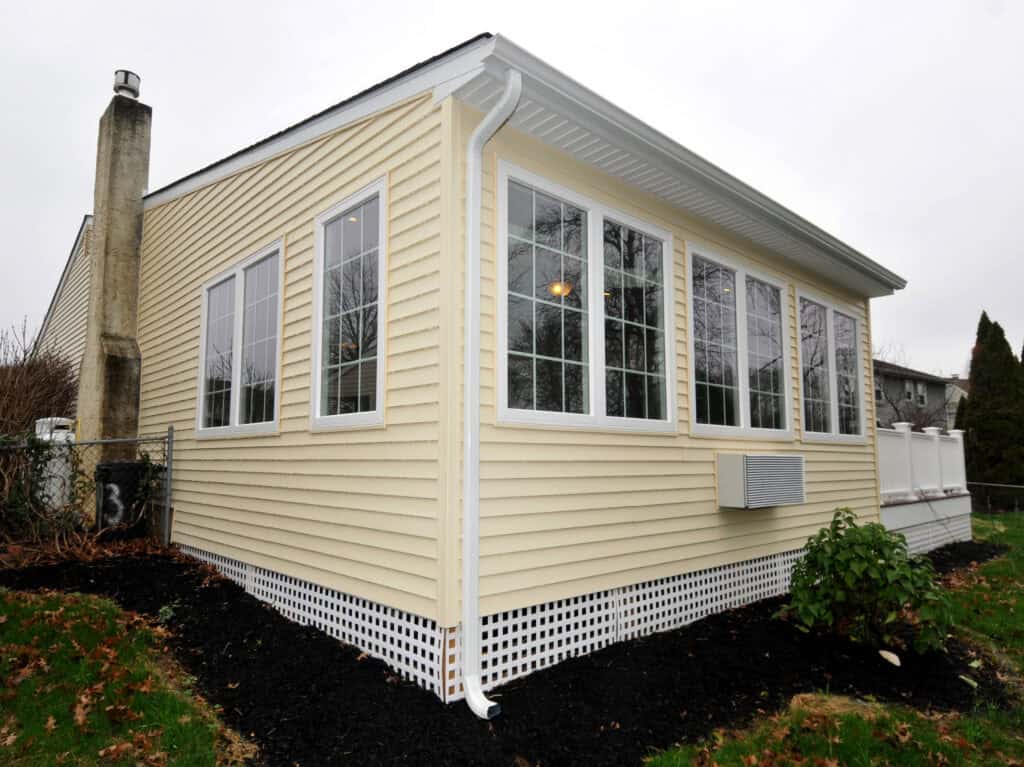 A yellow house with white trim and large windows, featuring a white gutter and lattice skirting at the foundation. The yard has mulch, sparse grass, and a few small shrubs, under a cloudy sky.