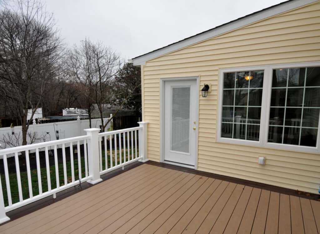 A beige house with white trim features a glass door and large windows opening onto a spacious, light brown and tan deck with white railings. Leafless trees and a white fence are visible in the background.