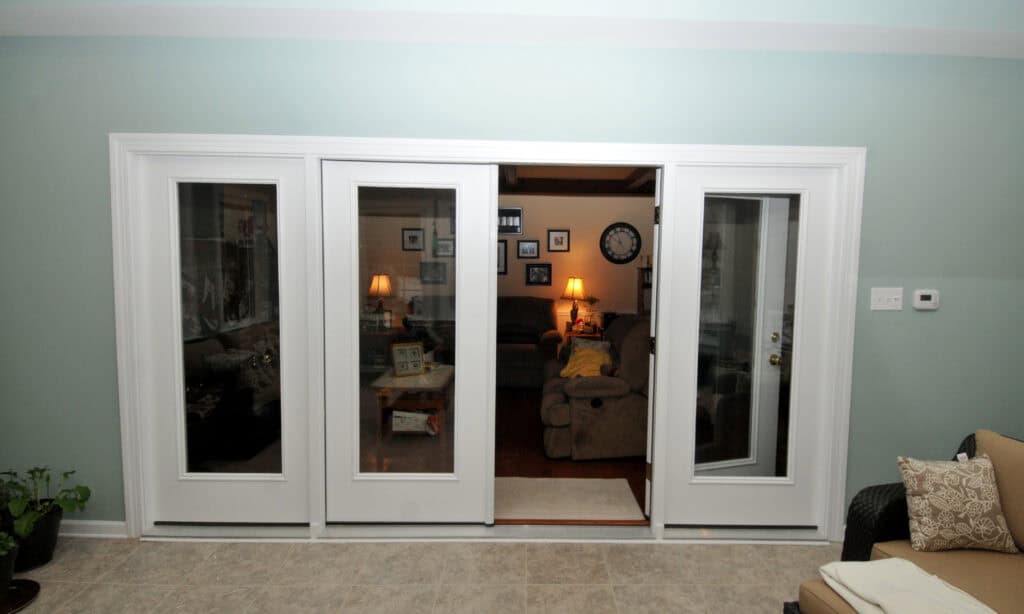 White French doors with large glass panes open into a cozy living room with brown sofas, soft lighting, framed photos on the walls, and a clock. The floor is tiled, and a plant sits in the left corner.