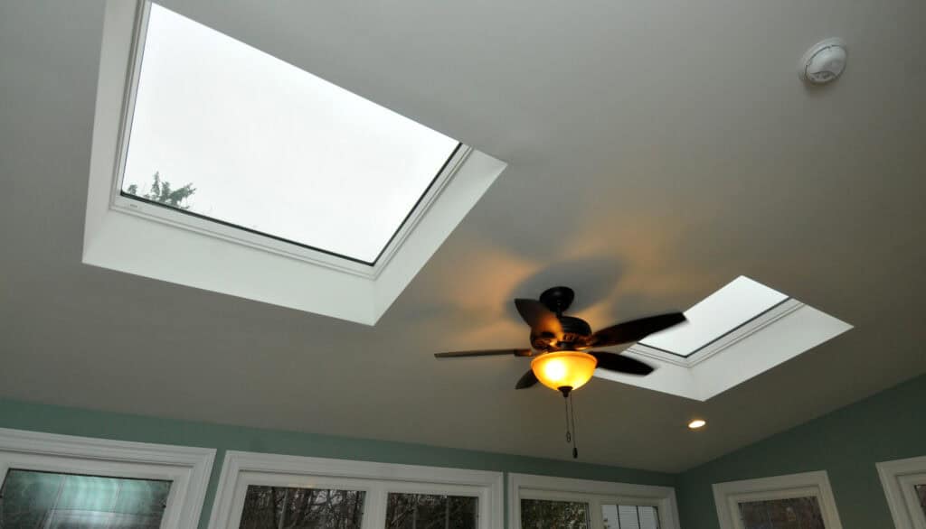 Two skylights in a white ceiling with a ceiling fan and light fixture in the center. The room has teal walls and large windows along one side.