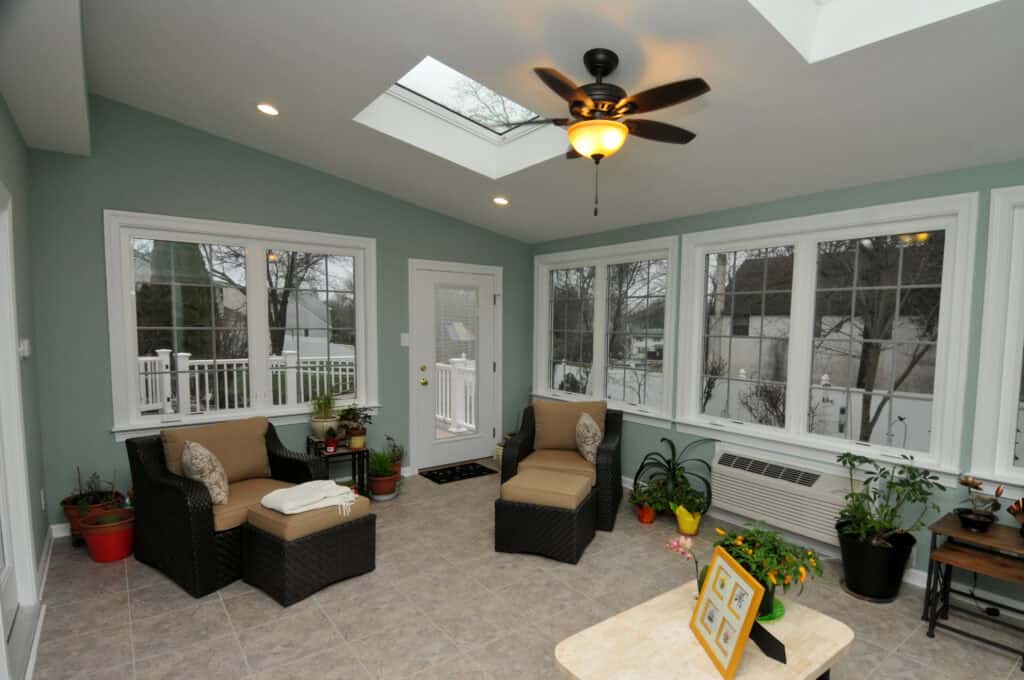 A bright sunroom with large windows, skylights, wicker chairs, potted plants, a ceiling fan, tiled floor, and a small table with framed photos and a yellow tablecloth.