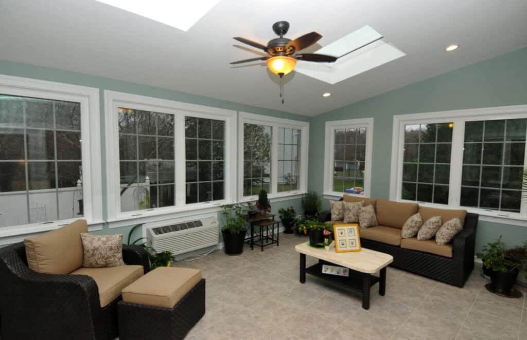 A bright sunroom with large windows, tiled floor, wicker furniture with beige cushions, potted plants, a coffee table, and a ceiling fan with a light. Skylights let in additional natural light.