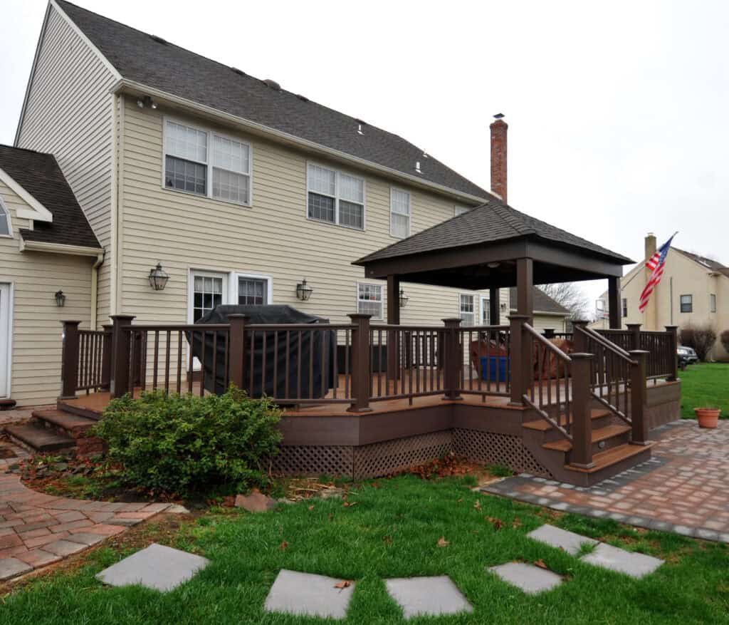 A raised wooden deck with railings and a covered seating area attached to the back of a beige two-story house, with steps leading down to a lawn and a stone path. An American flag and potted plant are nearby.