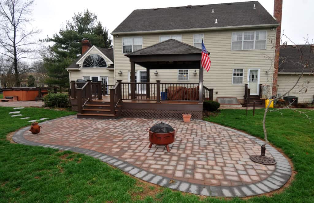 A backyard with a paved patio, fire pit, and a large wooden deck attached to a beige house. There is a covered area on the deck and an American flag hanging from a post. Surrounding the patio is green grass.