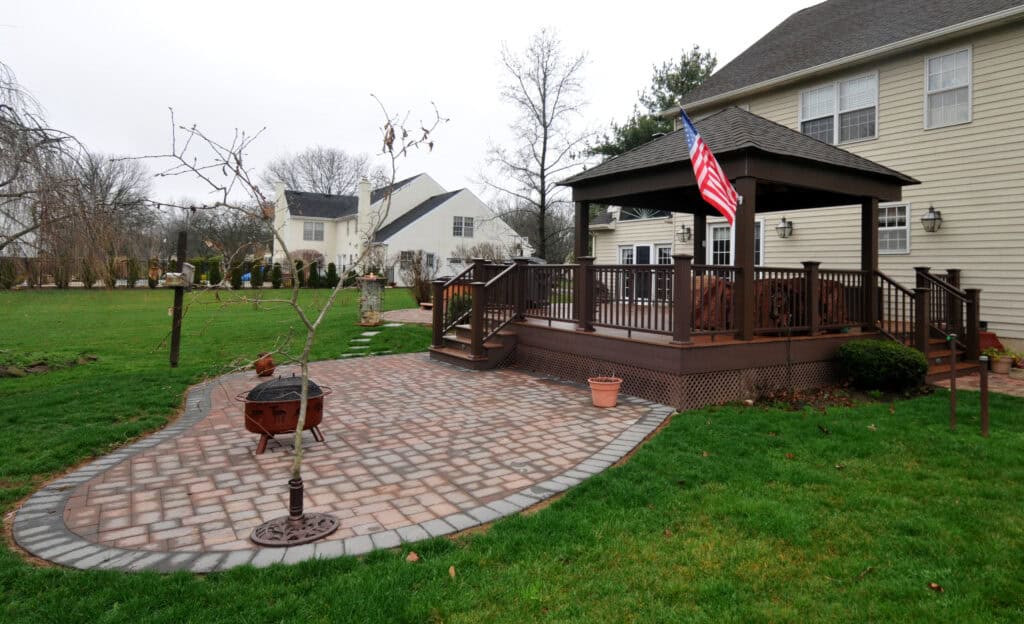 A backyard with a patio of interlocking bricks, a small tree, a fire pit, and a raised wooden deck with a covered section and an American flag. The yard has green grass and nearby houses.