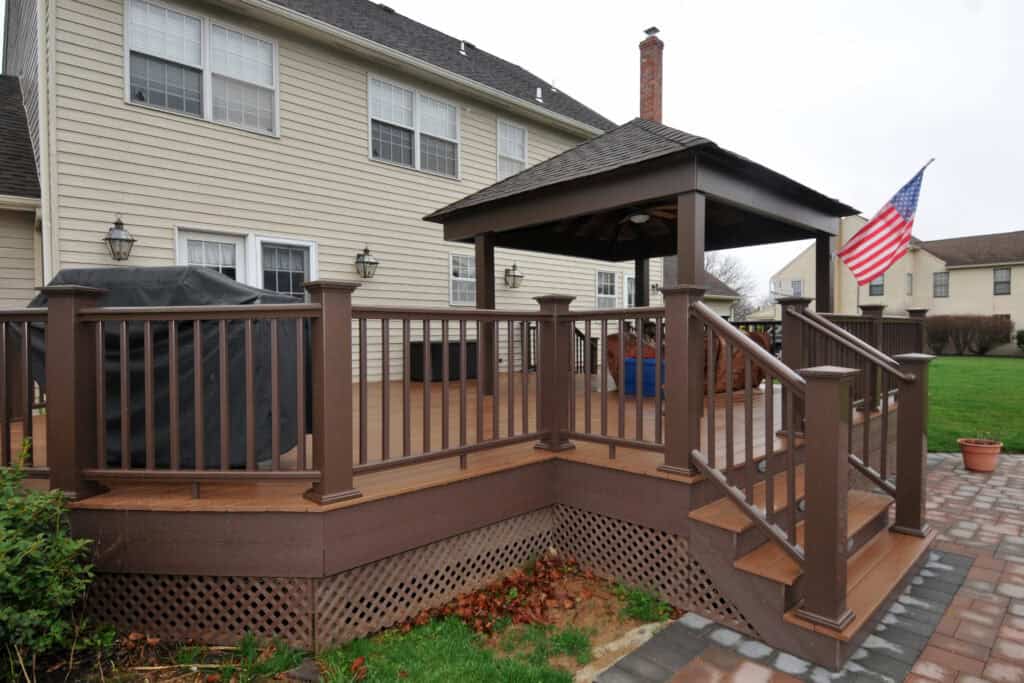 A brown wooden deck with stairs and railing, attached to a beige house. The deck features a grill, a covered gazebo area, and an American flag. The yard and paved patio are visible in the background.