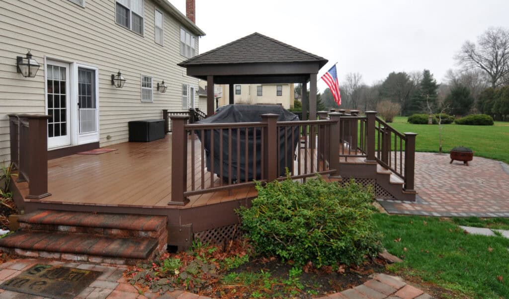 A brown wooden deck attached to a beige house features a covered gazebo, an outdoor grill with a cover, and an American flag. Brick steps and a walkway lead to a grassy yard with trees and a fire pit.