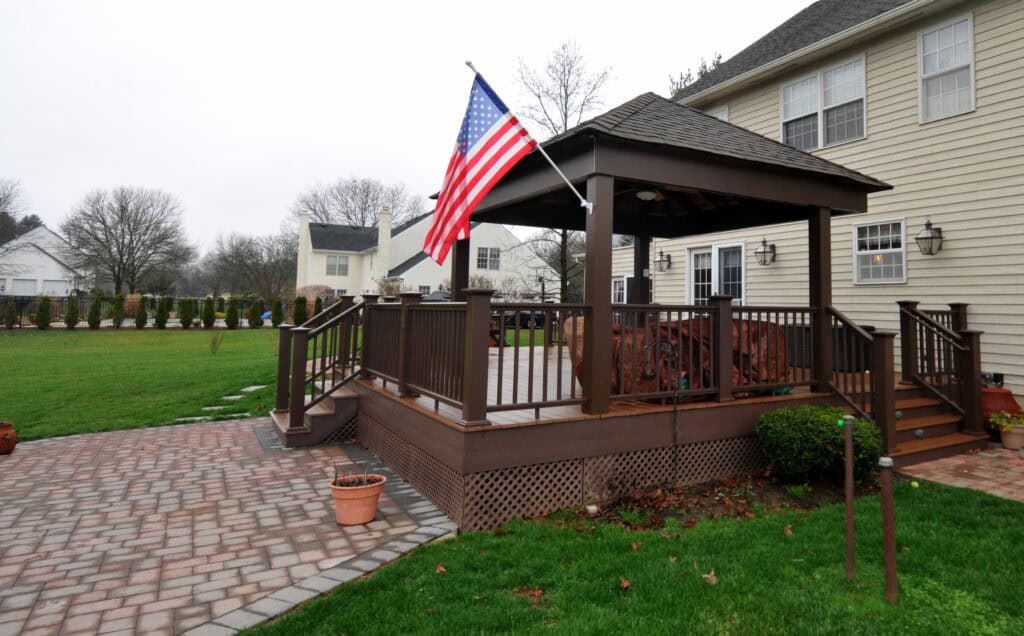 A wooden backyard gazebo with an American flag, surrounded by a deck and railing, sits next to a beige house with brick pathways, green lawn, and neighboring houses in the background.