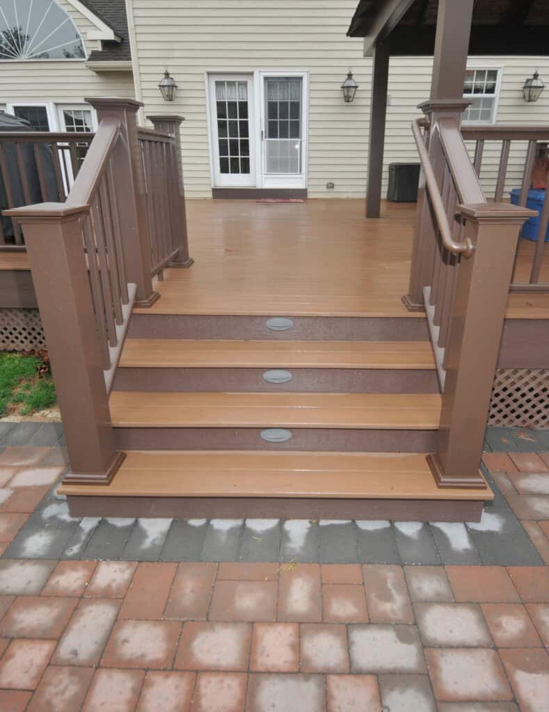 Brown wooden deck stairs with railings, leading up from a patterned patio to a covered deck attached to a beige house with white-trimmed windows and doors.