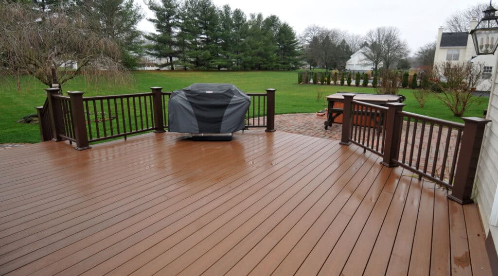 A spacious wooden deck with dark railings, featuring a covered grill in the center. The deck overlooks a large green backyard bordered by trees and a brick patio. The scene appears to be on a rainy or overcast day.