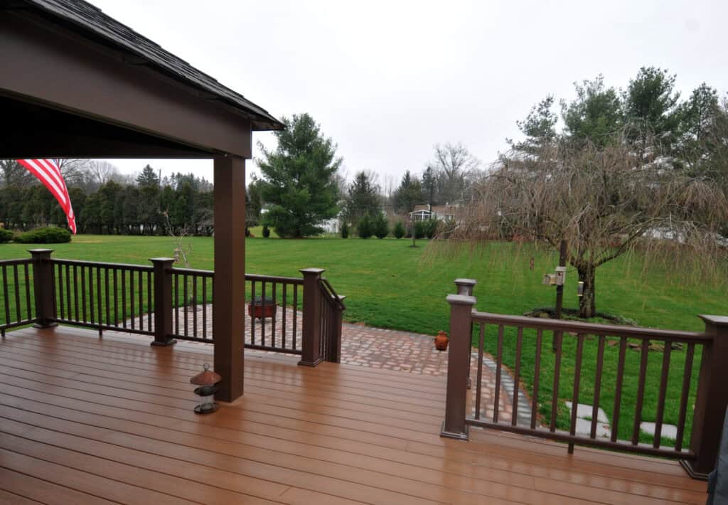 A view from a wooden deck overlooking a spacious, green backyard with trees, a brick patio area, and an American flag visible on the left side. The sky is overcast.