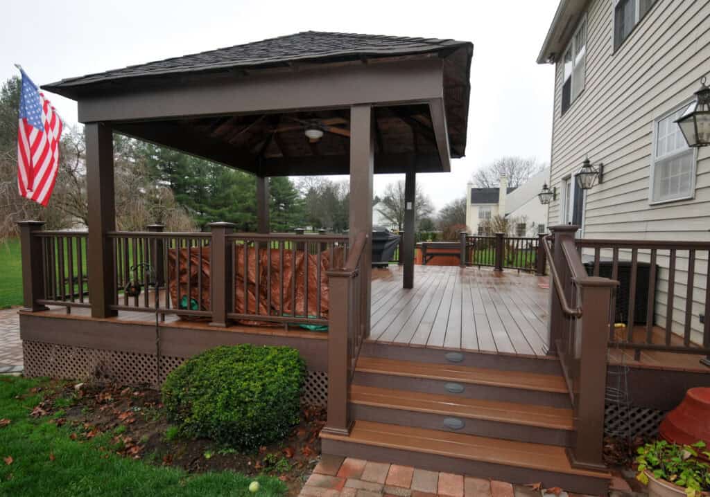 A wooden backyard deck with stairs, a gazebo, railings, and an American flag, attached to a beige house. The deck overlooks a grassy yard with bushes and trees.