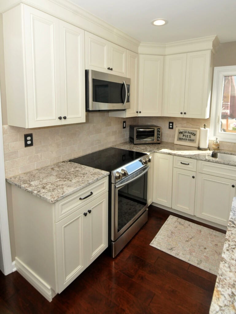 A modern kitchen with white cabinets, granite countertops, electric stove, microwave, small toaster oven, and dark wood floor. A window lets in natural light, and a decorative rug and sign complete the cozy space.