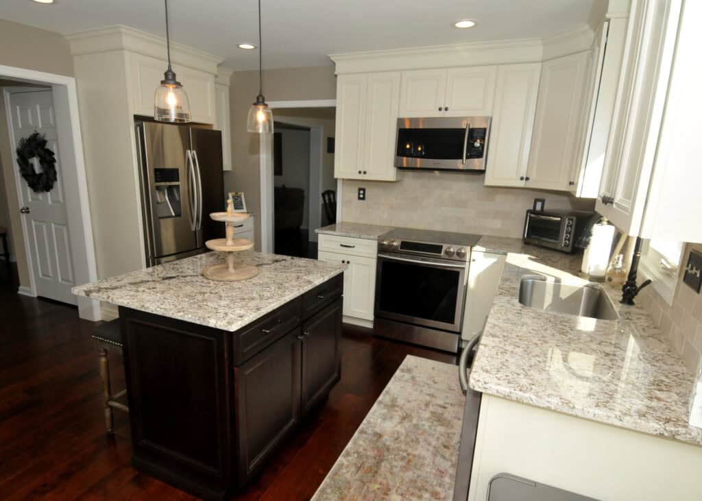 Modern kitchen with white cabinets, stainless steel appliances, a dark wood island, granite countertops, pendant lights, and a window above the sink; hardwood floors and a door with a wreath are also visible.