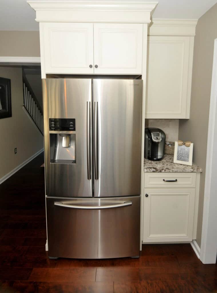 A modern stainless steel French door refrigerator with a water and ice dispenser is built into white cabinetry next to a granite countertop with a coffee maker and a small decorative tray. Dark hardwood floors.
