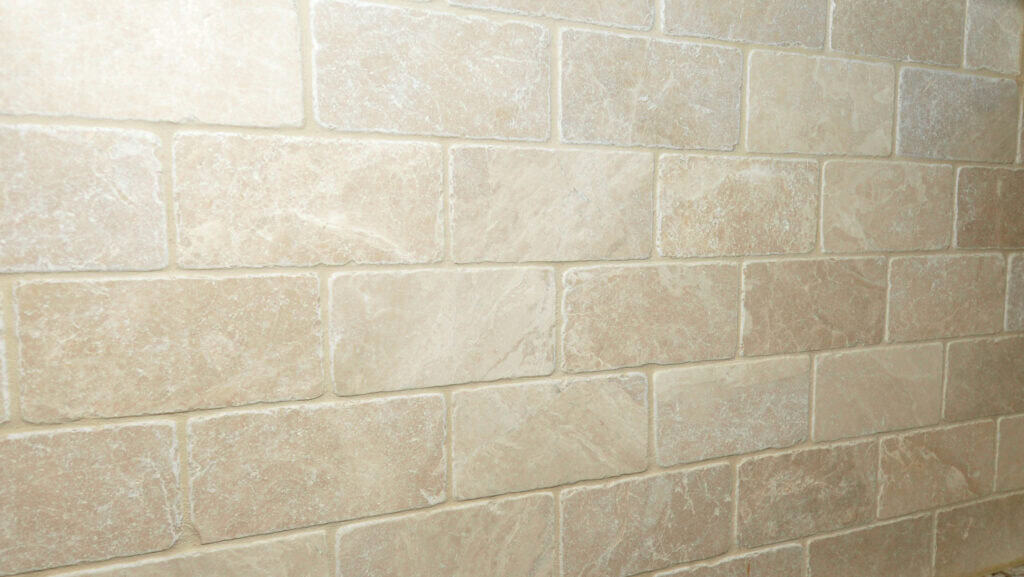A close-up view of a wall covered with rectangular beige stone tiles arranged in a staggered pattern, showing natural variations in color and texture.
