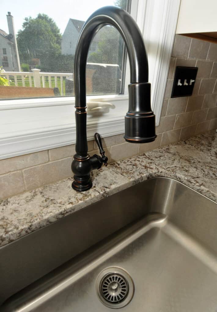 A modern dark bronze kitchen faucet with a curved neck is installed above a stainless steel sink on a speckled countertop, with a tiled backsplash and a window in the background.