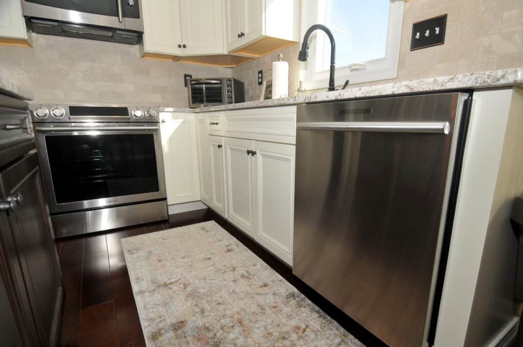 A modern kitchen with white cabinets, stainless steel appliances including a stove and dishwasher, a black faucet, a countertop with a paper towel holder, and a patterned rug on dark wood flooring.