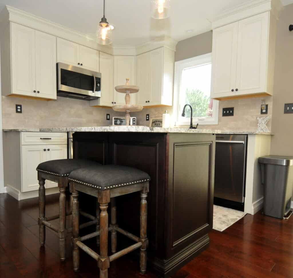 Modern kitchen with white cabinets, stainless steel appliances, a dark wood island with two cushioned stools, granite countertops, pendant lights, and a window above the sink.
