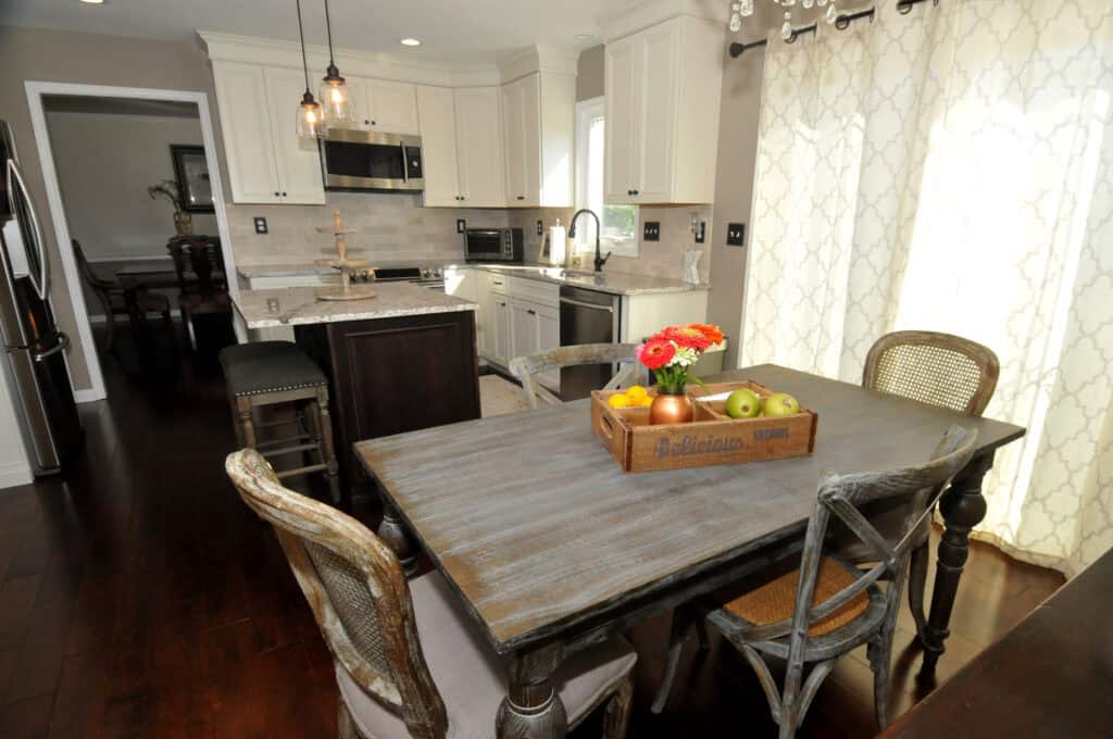 Bright kitchen with white cabinets, stainless steel appliances, a central island with stools, and a rustic dining table holding a wooden tray with fruit and flowers near patterned curtains.