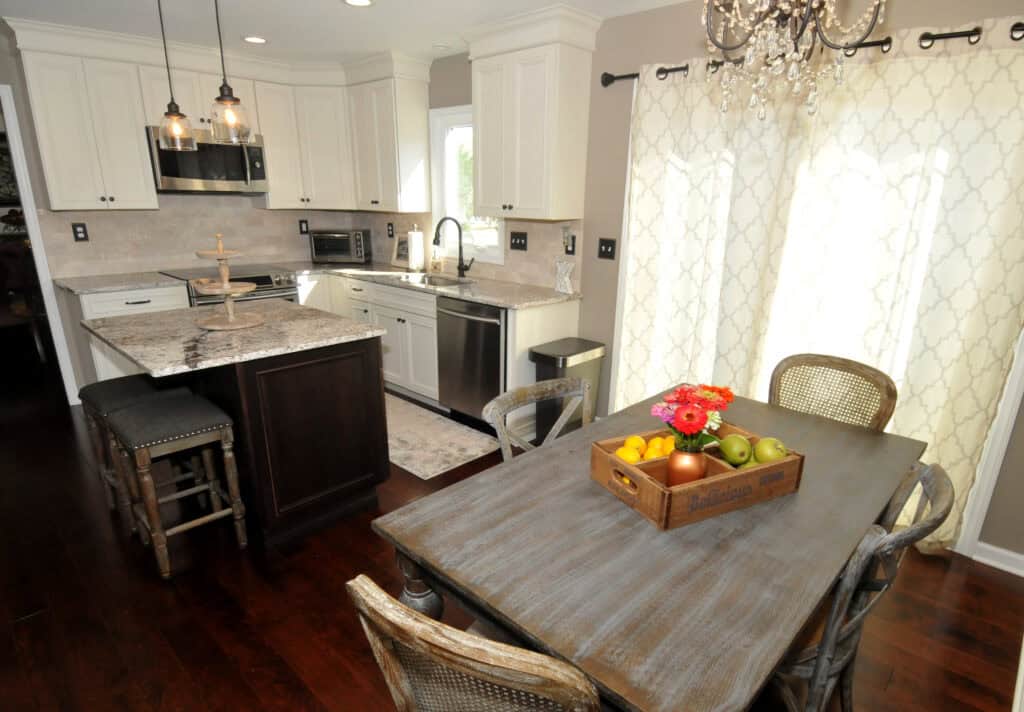 Bright kitchen and dining area with white cabinets, granite countertops, stainless steel appliances, and a wooden table set with a tray of fruit and flowers. Pendant lights and a chandelier add elegance to the space.
