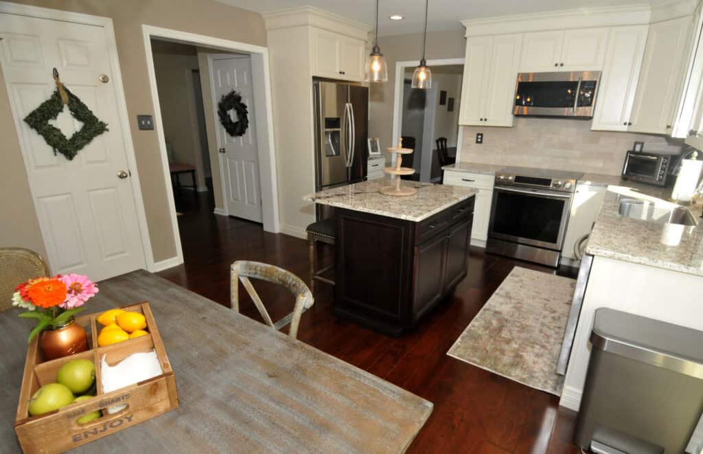 A modern kitchen with white cabinets, stainless steel appliances, a dark wood island, and granite countertops. A dining table with a crate of fruit and flowers is in the foreground; two doors have wreaths hanging on them.