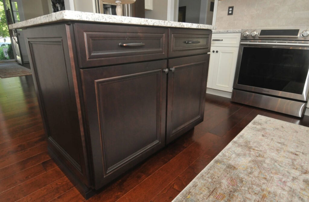 A close-up view of a dark wood kitchen island with a marble countertop, situated on a polished wood floor near a stove and white cabinetry.