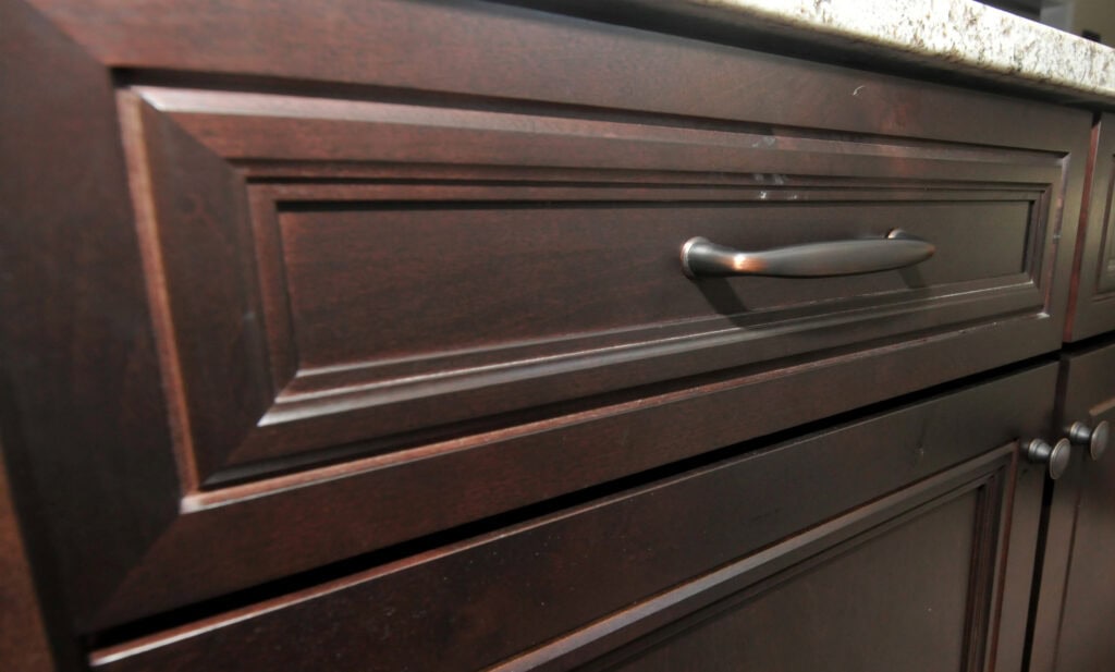 Close-up of a dark brown wooden kitchen cabinet drawer with a silver handle and part of a granite countertop visible at the top.