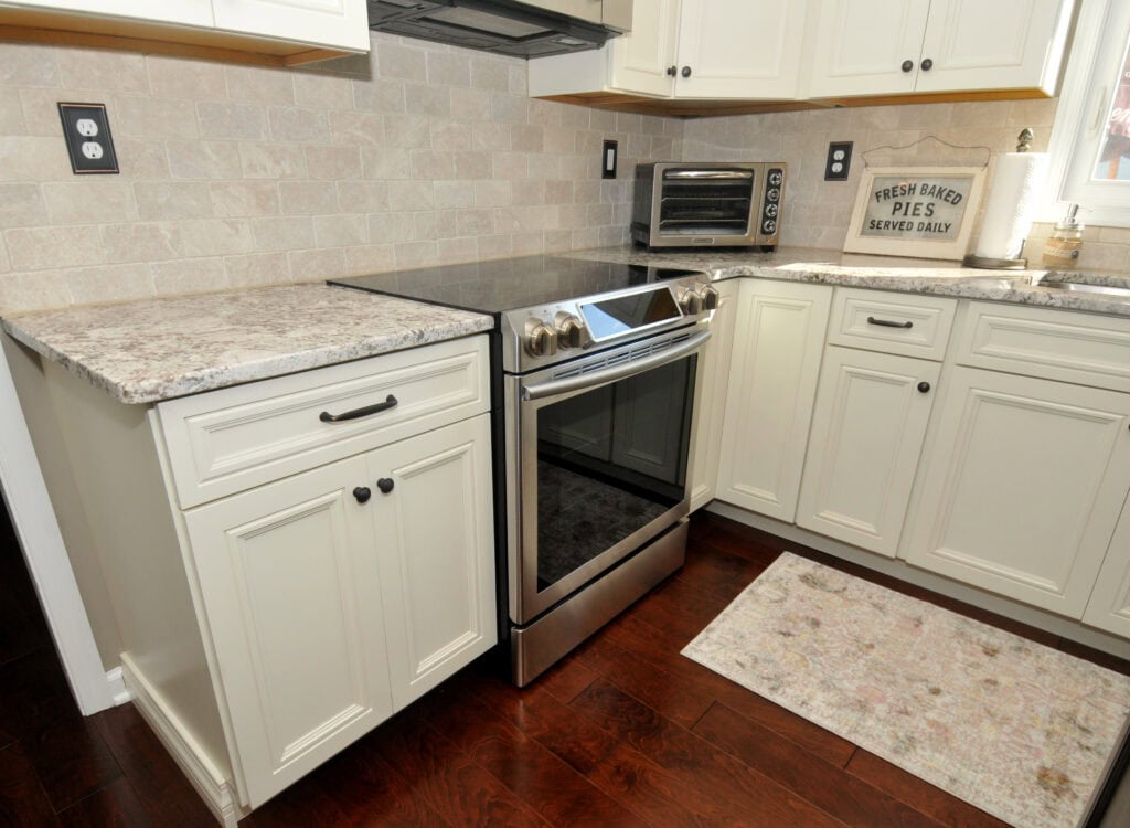A modern kitchen with white cabinets, granite countertops, a stainless steel electric stove, a small toaster oven, and a beige rug on dark wood flooring. A sign on the counter reads Fresh Baked Pies Served Daily.