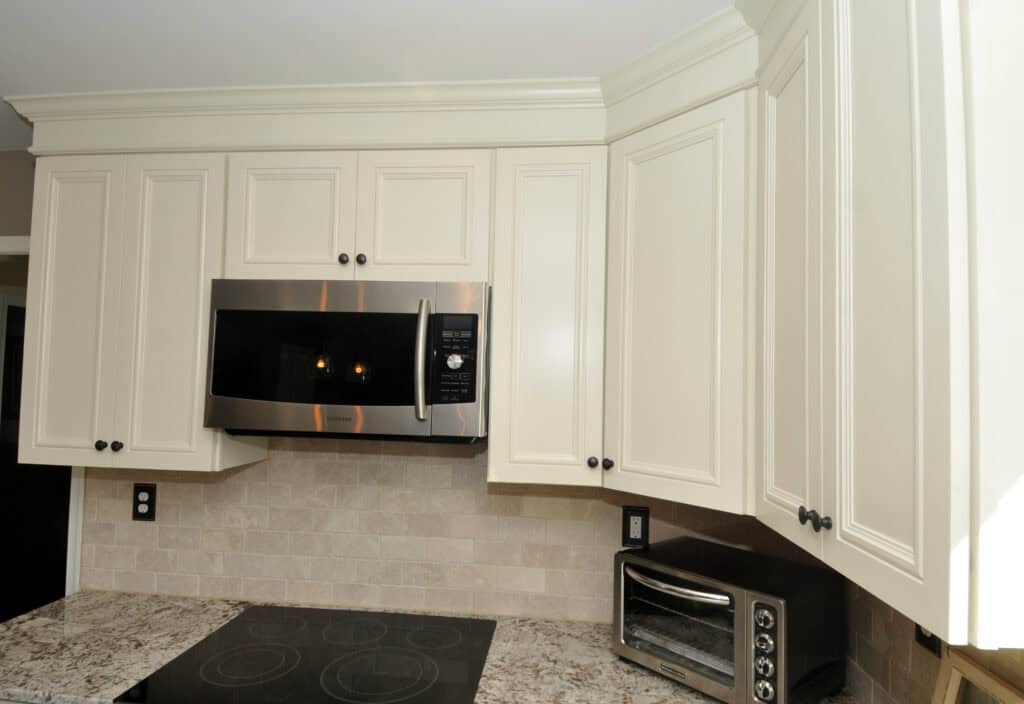 A modern kitchen features cream-colored cabinets, a stainless steel microwave, a small toaster oven, and a smooth electric cooktop on a granite countertop, with light beige subway tile backsplash.