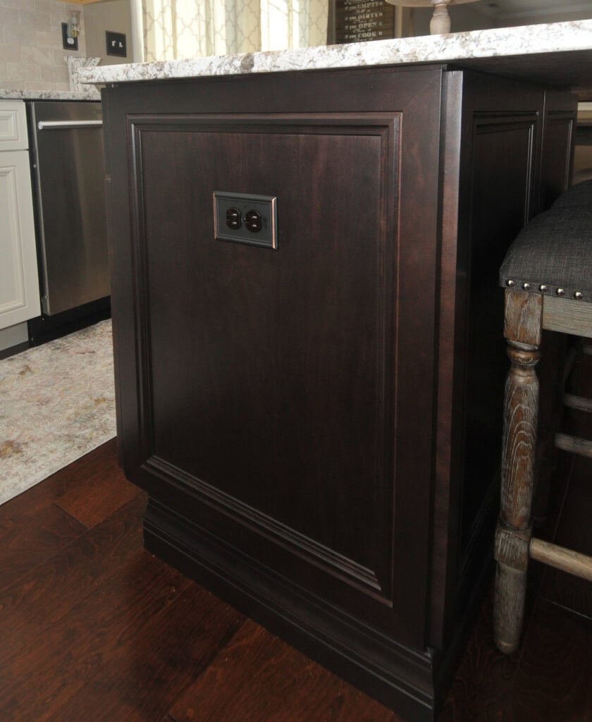 A kitchen island with dark wood paneling features a double electrical outlet on the side. Marble countertop, gray upholstered stool, and part of a stainless steel dishwasher are visible.