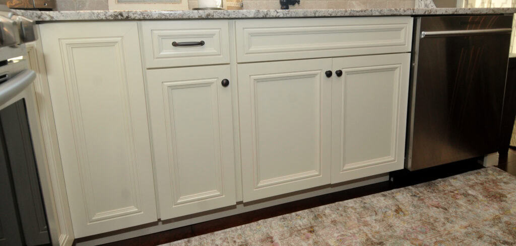 White kitchen cabinets with black knobs and a single drawer handle, next to a stainless steel dishwasher. The cabinets sit below a light-colored countertop, and a patterned rug is partially visible on the floor.