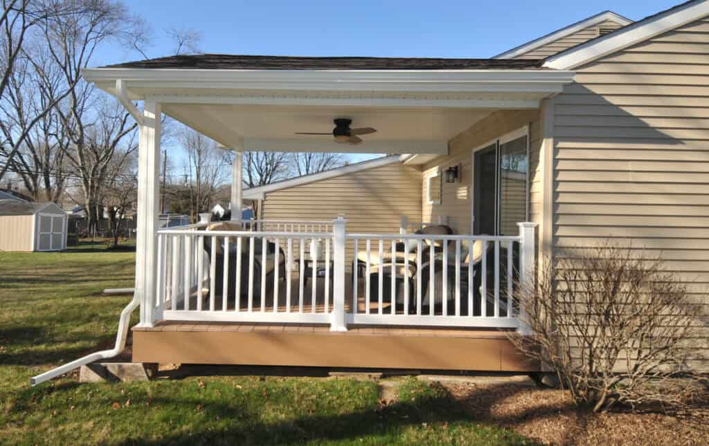 A covered backyard porch attached to a beige house, featuring white railings, a ceiling fan, outdoor seating, and a grassy yard with bare trees and a shed in the background.