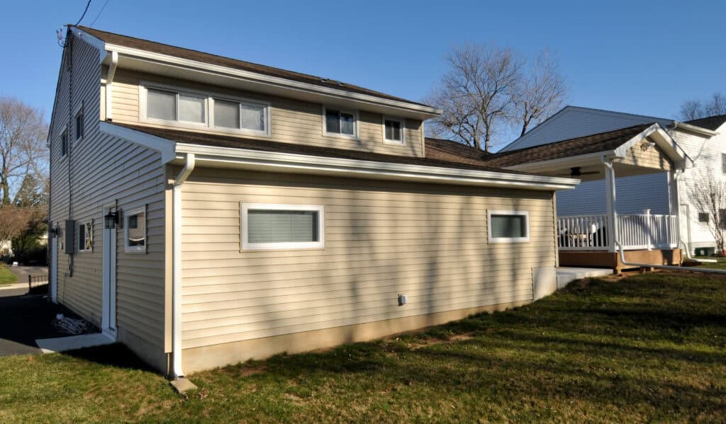 A beige, two-story house with horizontal siding, small rectangular windows, and a sloped roof, sits on a grassy lawn under a clear blue sky. Another house is partially visible next door.