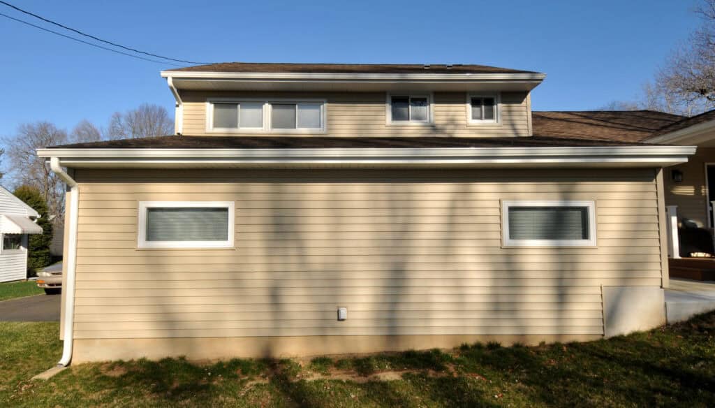 A beige house with horizontal siding, two rectangular windows on the lower level, and several smaller windows on an upper level. The lawn is green and tree shadows fall across the wall on a clear, sunny day.