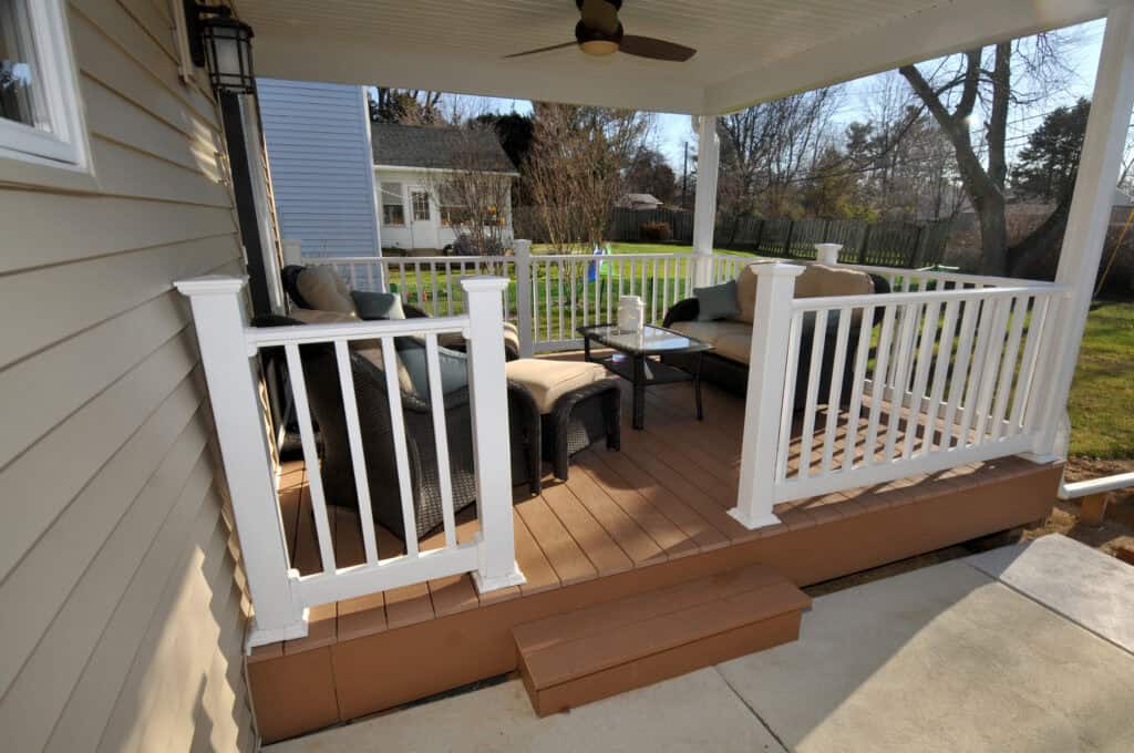 A cozy porch with wicker chairs, a loveseat, cushions, and a glass-top table sits behind white railings. There is a ceiling fan above and steps leading down to a sunny, grassy backyard.