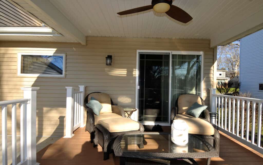 A cozy porch with two cushioned wicker chairs, a glass coffee table, and a ceiling fan. The porch has white railings, beige siding, and a sliding glass door leading inside. Sunlight streams across the space.