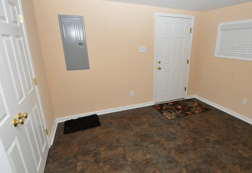 A small room with beige walls, a white door, a window with closed blinds, a wall-mounted electrical panel, two rugs on a brown tile floor, and white double doors on the left.