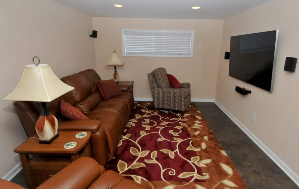 A cozy home theater room with brown leather couches, a patterned armchair, a red and beige floral rug, lamps, an end table, and a flat-screen TV mounted on the wall.