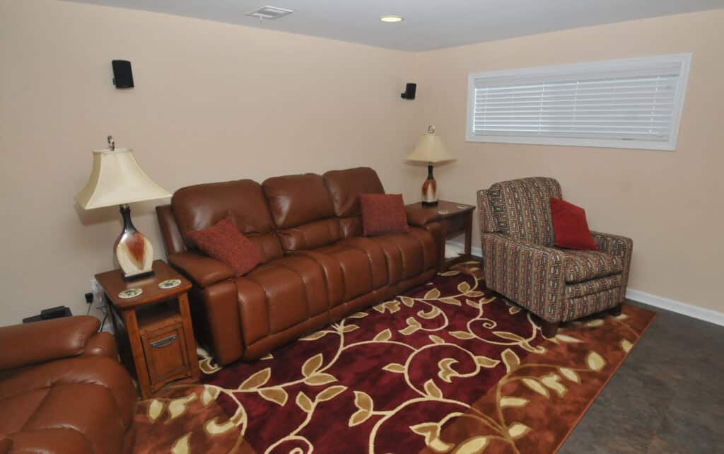 A cozy living room with a brown leather sofa, patterned armchair, two side tables with lamps, and a red and beige floral area rug. A window with white blinds is on the wall.