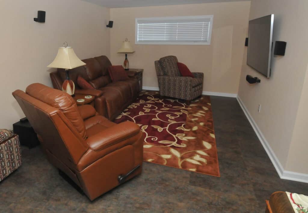 A cozy living room with brown leather recliners, a patterned armchair, a sofa, two lamps, a patterned red and beige rug, and a wall-mounted TV under a window with closed blinds.