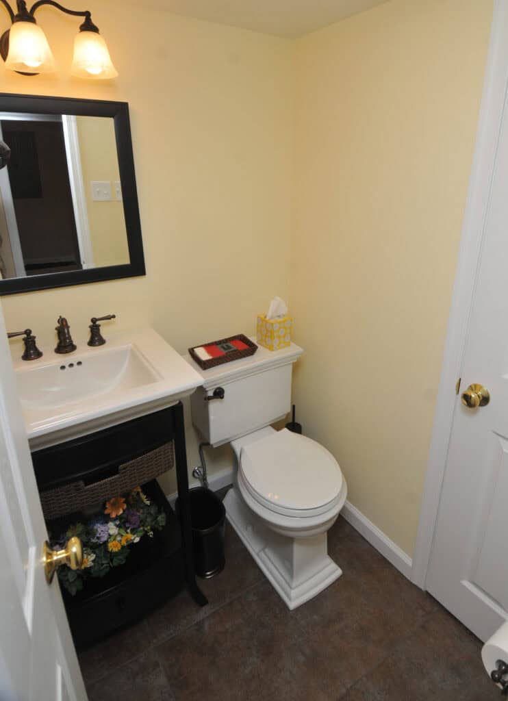 A small bathroom with a white toilet next to a white sink vanity, a black framed mirror above the sink, a tissue box and books on the toilet tank, yellow walls, and brown tiled floor.