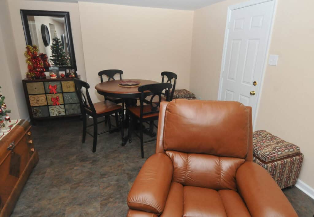 A cozy room with a brown leather recliner in the foreground, a round wooden dining table with four chairs, a mirror, and holiday decorations, including a small Christmas tree on a cabinet.