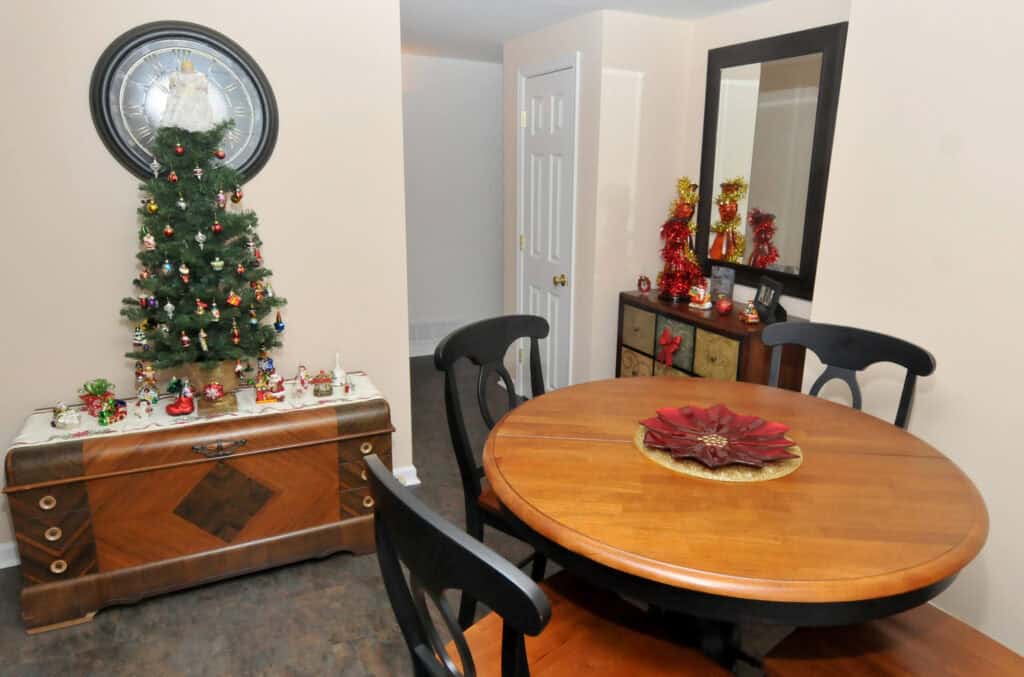 A small dining room decorated for Christmas, featuring a round wooden table with four chairs, a poinsettia centerpiece, a chest with a small decorated Christmas tree, and a mirror with festive decor on a dresser.
