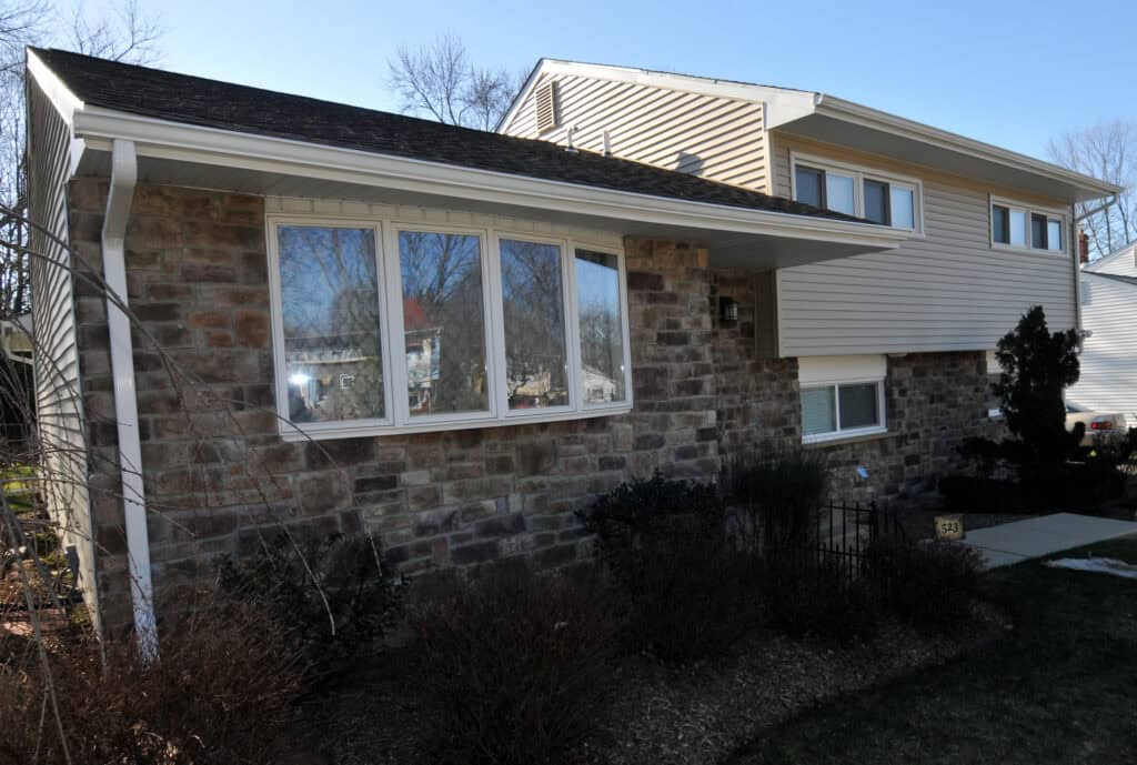 A split-level house with stone and beige siding, large front windows, and a small garden with leafless bushes, photographed on a sunny day.