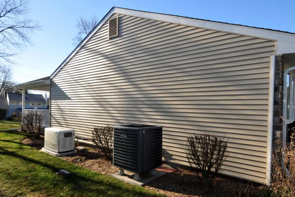 A beige house exterior with vinyl siding features an air conditioning unit and a backup generator on the ground, surrounded by small bushes and a grassy lawn, under a clear blue sky.