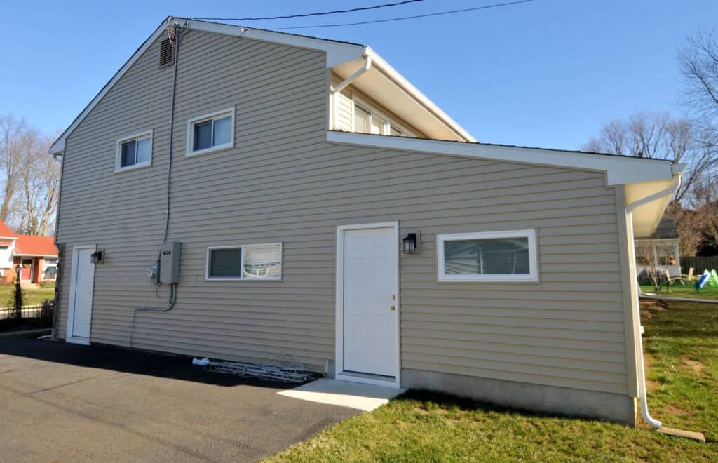 A beige two-story house with horizontal siding, white-framed windows, a white door, and a sloped driveway. Electrical cords are coiled near the foundation. The sky is clear and the yard is partially visible.
