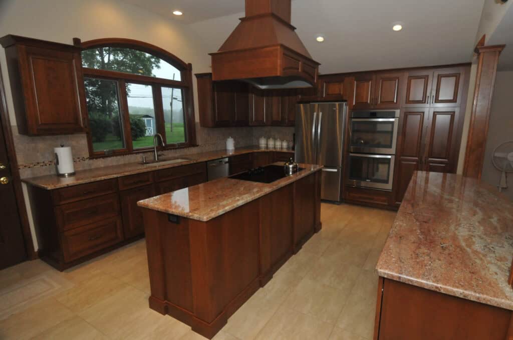 Spacious kitchen with dark wood cabinets, granite countertops, stainless steel appliances, an island in the center, large windows, and beige tiled flooring.
